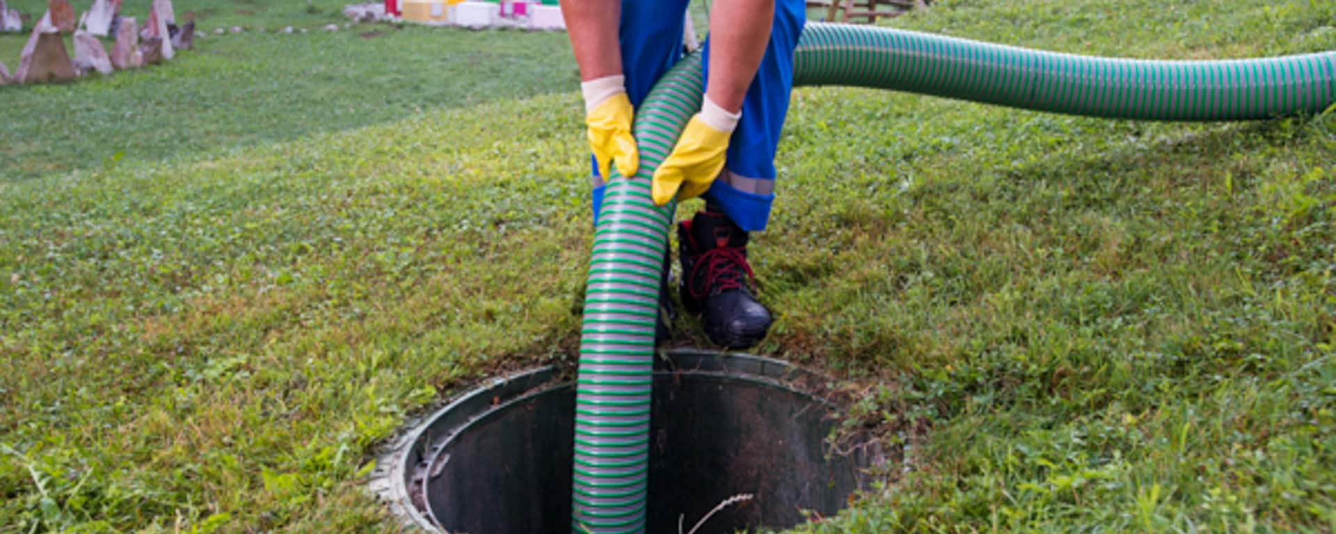 Plumbing technician handling a green striped hose over a sewer access point in a grassy yard, illustrating drain maintenance services.