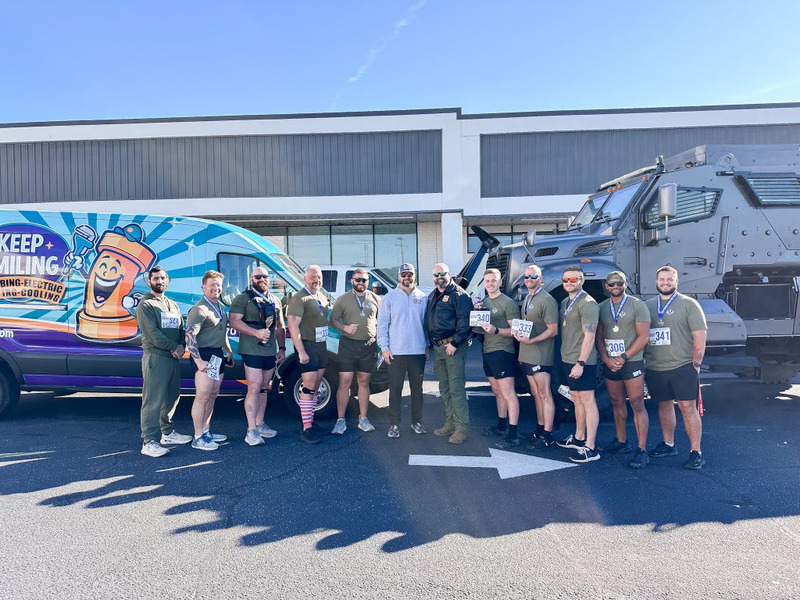 Team members of Keep Smiling Plumbing Electric Heating and Cooling posing with medals after participating in the SWAT Trot event, alongside a branded service van and police vehicle, showcasing community involvement and teamwork.