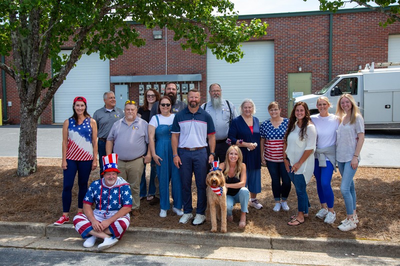 Group of Keep Smiling team members celebrating Independence Day at the office July 4th cookout, wearing patriotic attire, with a dog in festive gear, in front of a brick building.