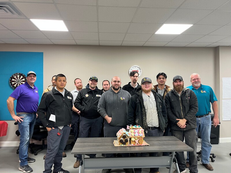 Group of Keep Smiling Plumbing Electric Heating and Cooling technicians posing together with a decorated gingerbread house during a holiday contest, showcasing community spirit and teamwork in a festive office setting.