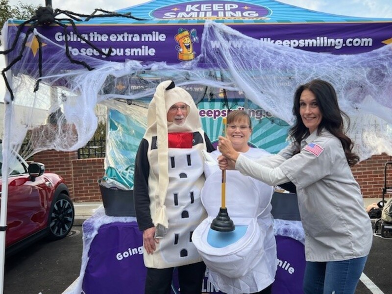 Technicians in Halloween costumes, including a snowman and a toilet, posing with a plunger under a Keep Smiling tent decorated for the holiday, showcasing community engagement and team spirit.