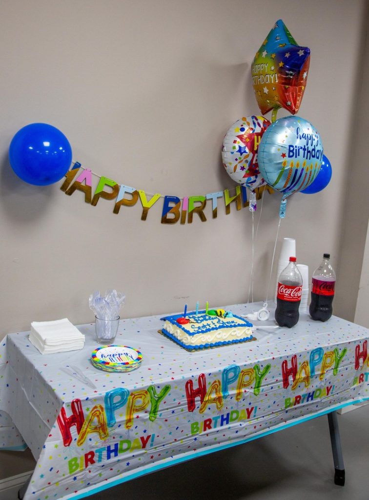 Birthday celebration setup with a decorated table featuring a cake, "Happy Birthday" banner, colorful balloons, and refreshments, highlighting team camaraderie at Keep Smiling.