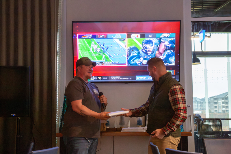 Technician presenting an award during a holiday party, with a sports game displayed on a screen in the background, reflecting team spirit and celebration.