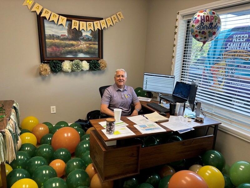 General Manager Jim celebrating his birthday surrounded by colorful balloons and a "Happy Birthday" banner in the Keep Smiling office.