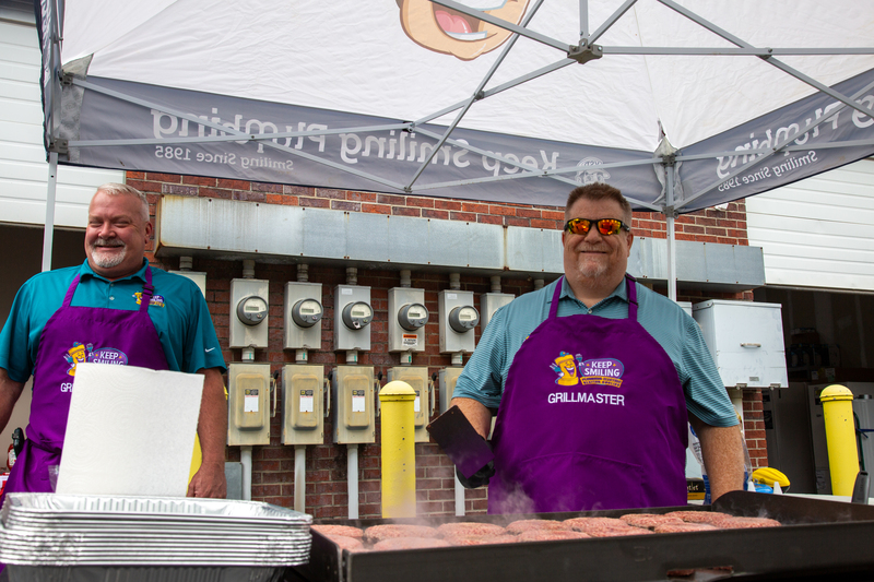 Team members grilling at a Memorial Day cookout, wearing "Keep Smiling" aprons, with a banner overhead, showcasing community spirit and teamwork.