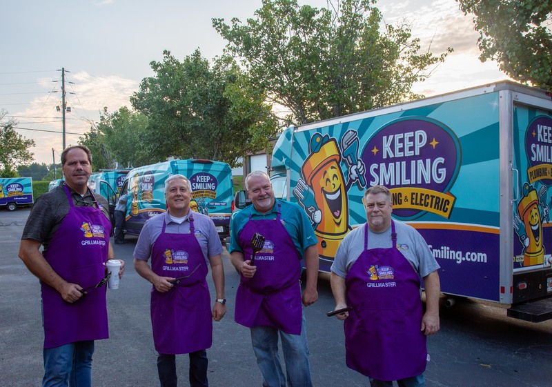 Four team members in purple aprons labeled "GRILLMASTER" smiling at a community event, with "Keep Smiling Plumbing & Electric" service vehicles in the background, showcasing team camaraderie and community engagement.