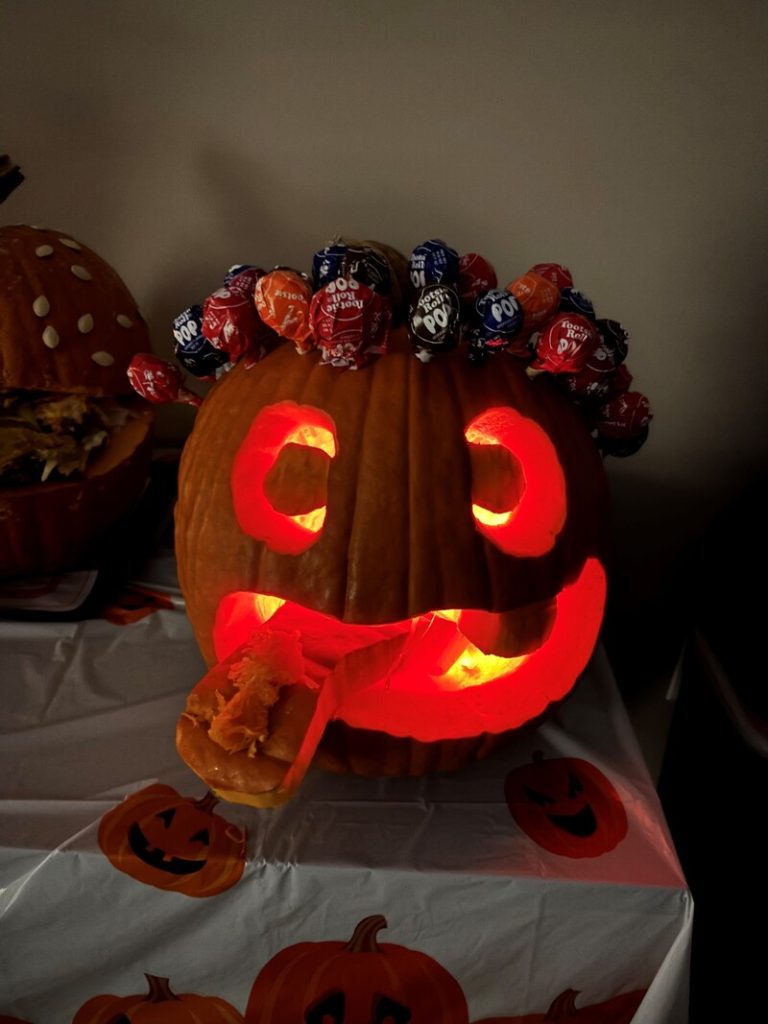 Halloween-themed carved pumpkin with glowing eyes and a mouth, adorned with colorful lollipops on top, and a piece of pumpkin sticking out, set against a festive tablecloth.